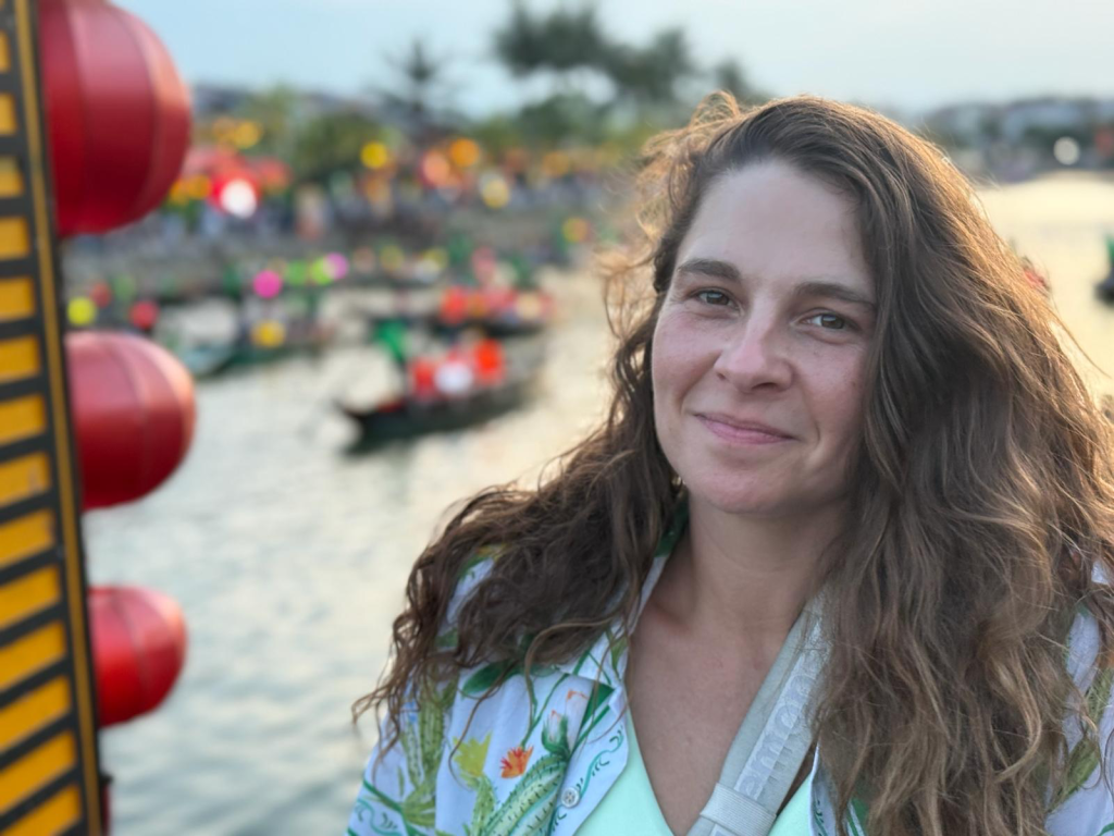 A person with long, wavy hair standing by a waterway, smiling lightly. In the background, colorful lanterns and boats filled with people can be seen.